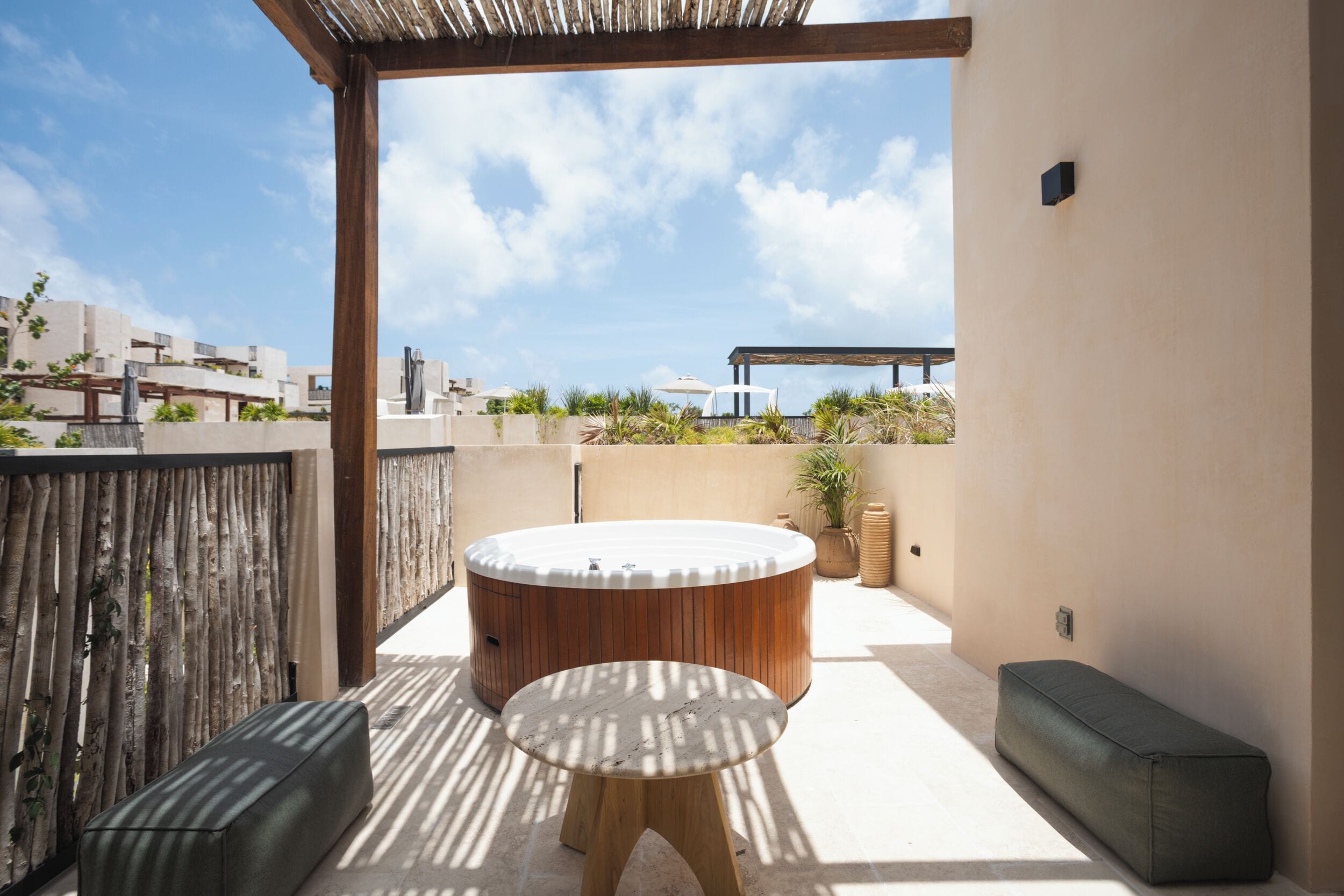An outdoor patio with a round wooden table and two stools, a pergola, and a view of the cityscape.