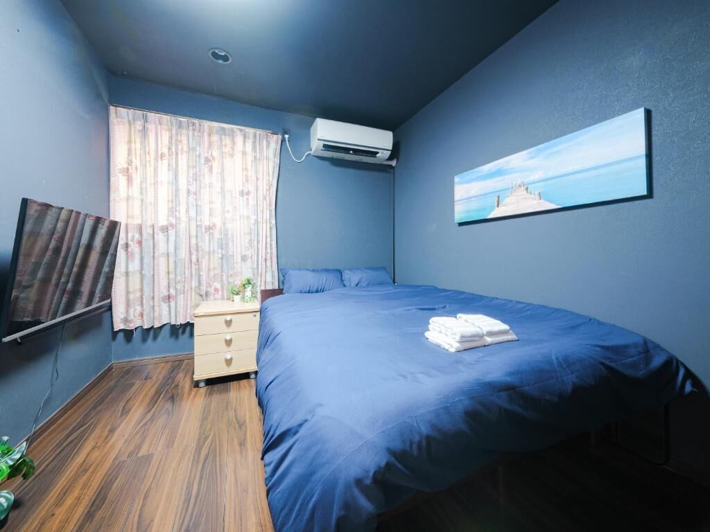 A bedroom with a bed, a nightstand, a wall-mounted air conditioner, a window with curtains, and a painting on the wall.