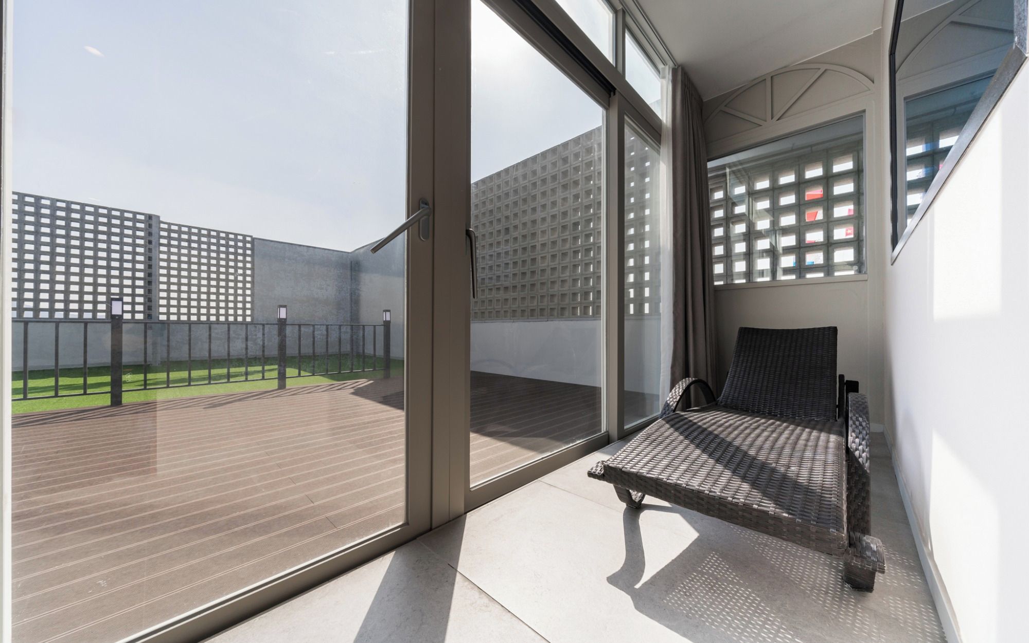 Un balcon donnant sur un immeuble à la façade quadrillée, avec une porte-fenêtre coulissante et un fauteuil en osier.