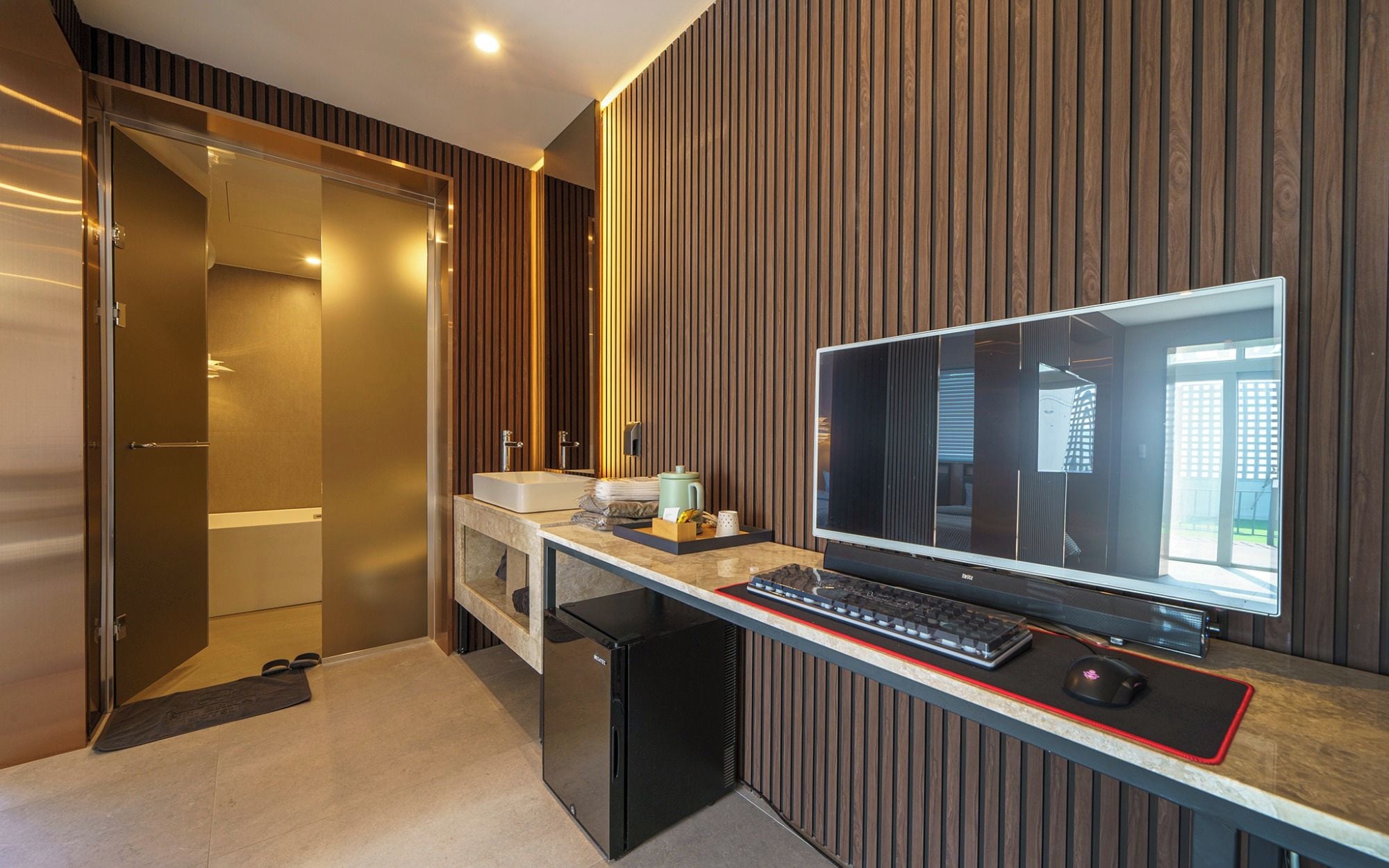 A modern bathroom with a shower, a sink, and a toilet. There is a computer desk with a monitor, keyboard, and mouse.
