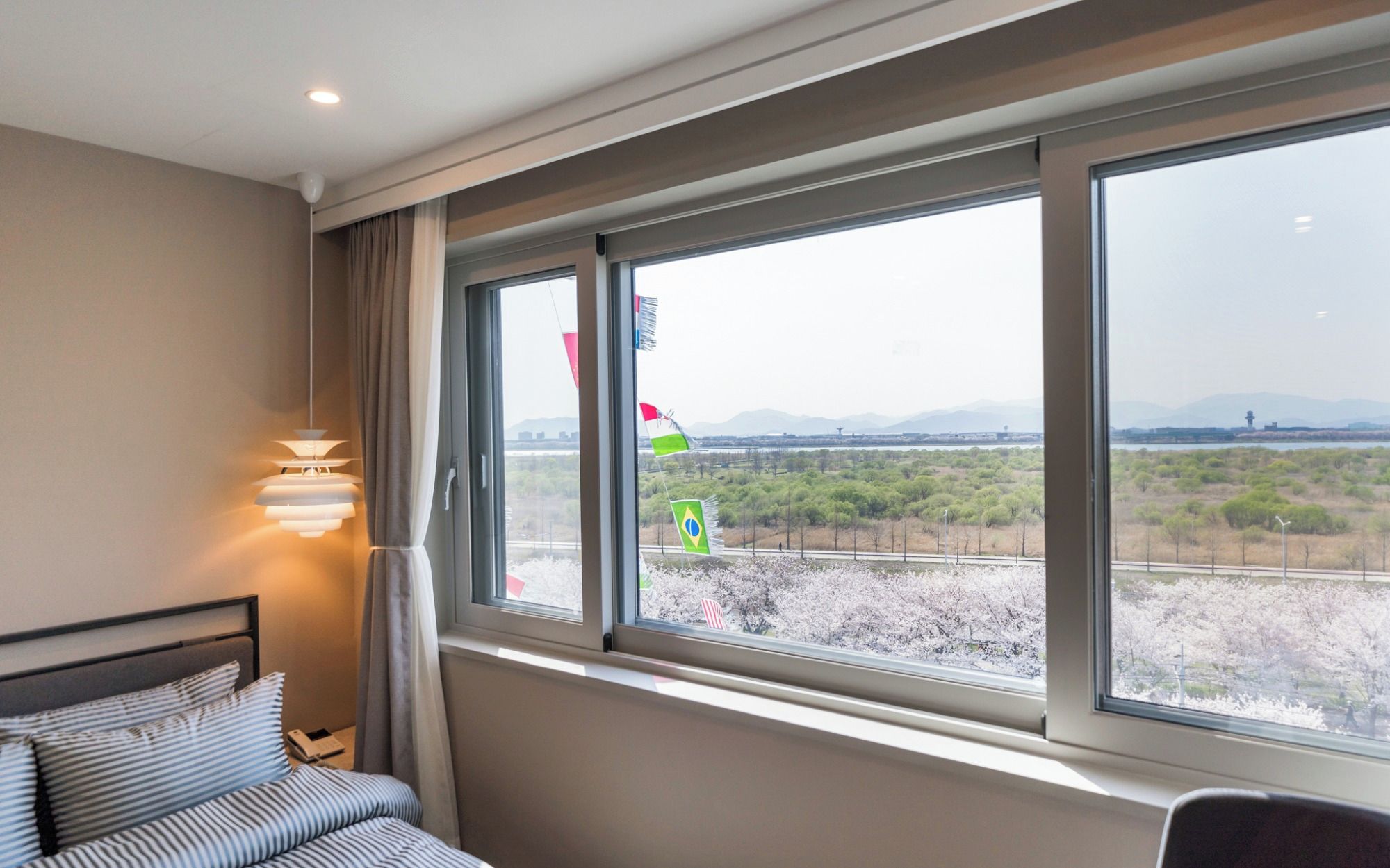 A hotel room with a bed, bedside table, lamp, and a large window offering a view of a landscape with trees and mountains.