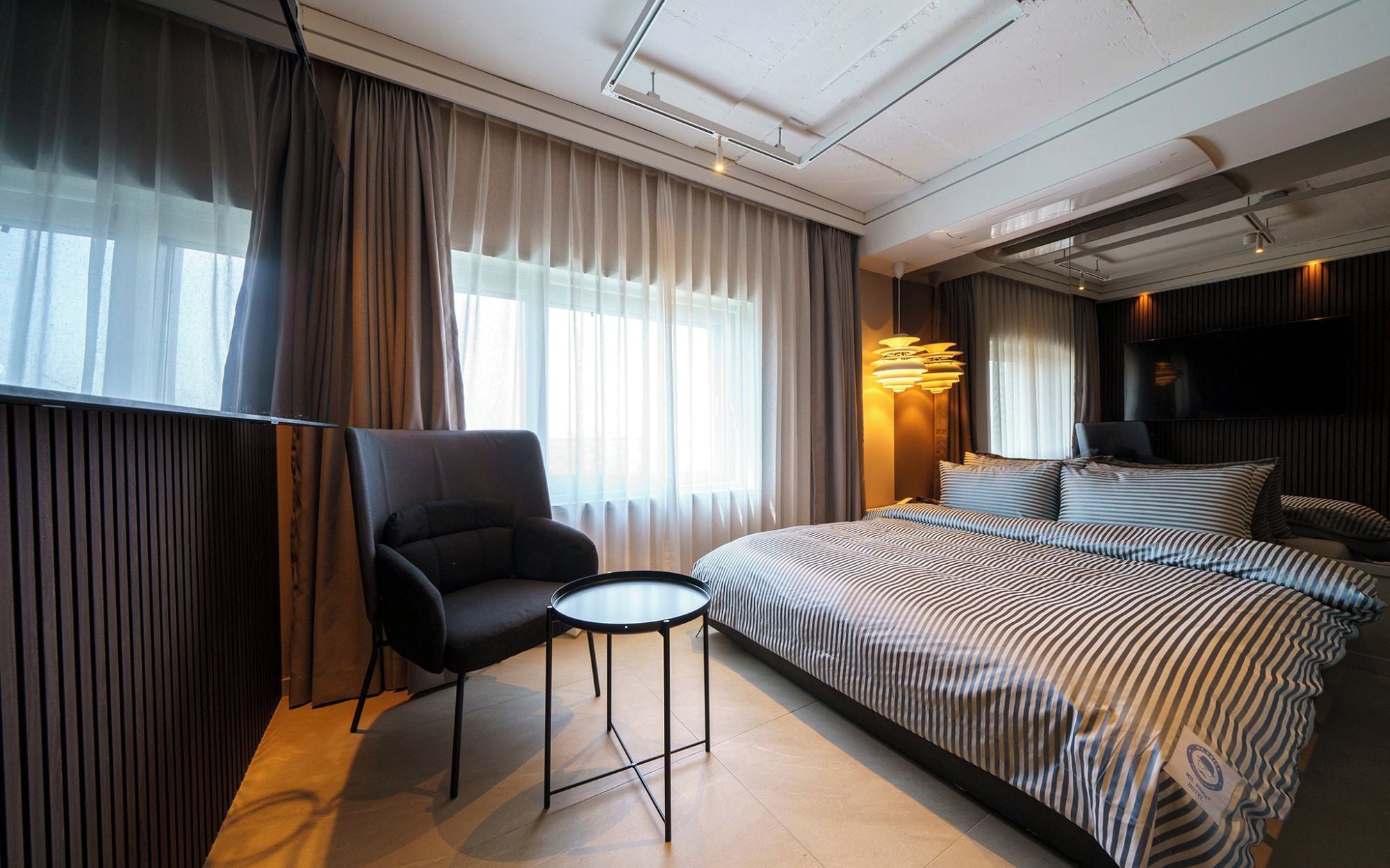 A hotel room with a bed, a chair, a small table, a TV, and a window with curtains.