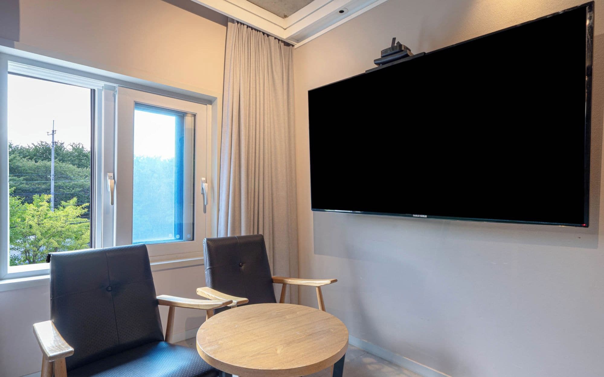 Une chambre d’hôtel dotée d’une grande fenêtre, de deux fauteuils, d’une table ronde et d’un téléviseur à écran plat fixé au mur.