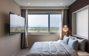 Habitación de hotel con cama, televisor montado en la pared, ventana con vista a un paisaje desértico y lámpara en la mesita de noche.