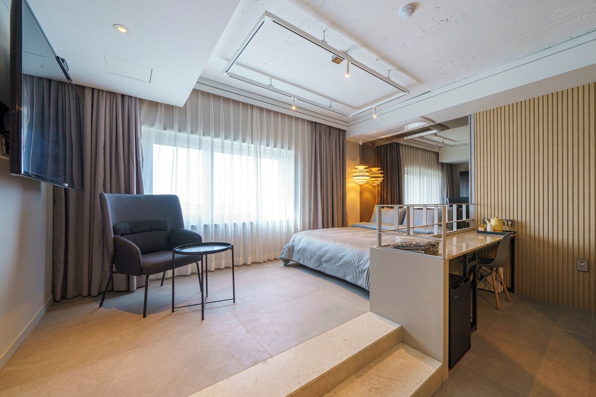 Ein Hotelzimmer mit Bett, Schreibtisch, Sessel, Fernseher und einem großen Fenster mit Vorhängen.