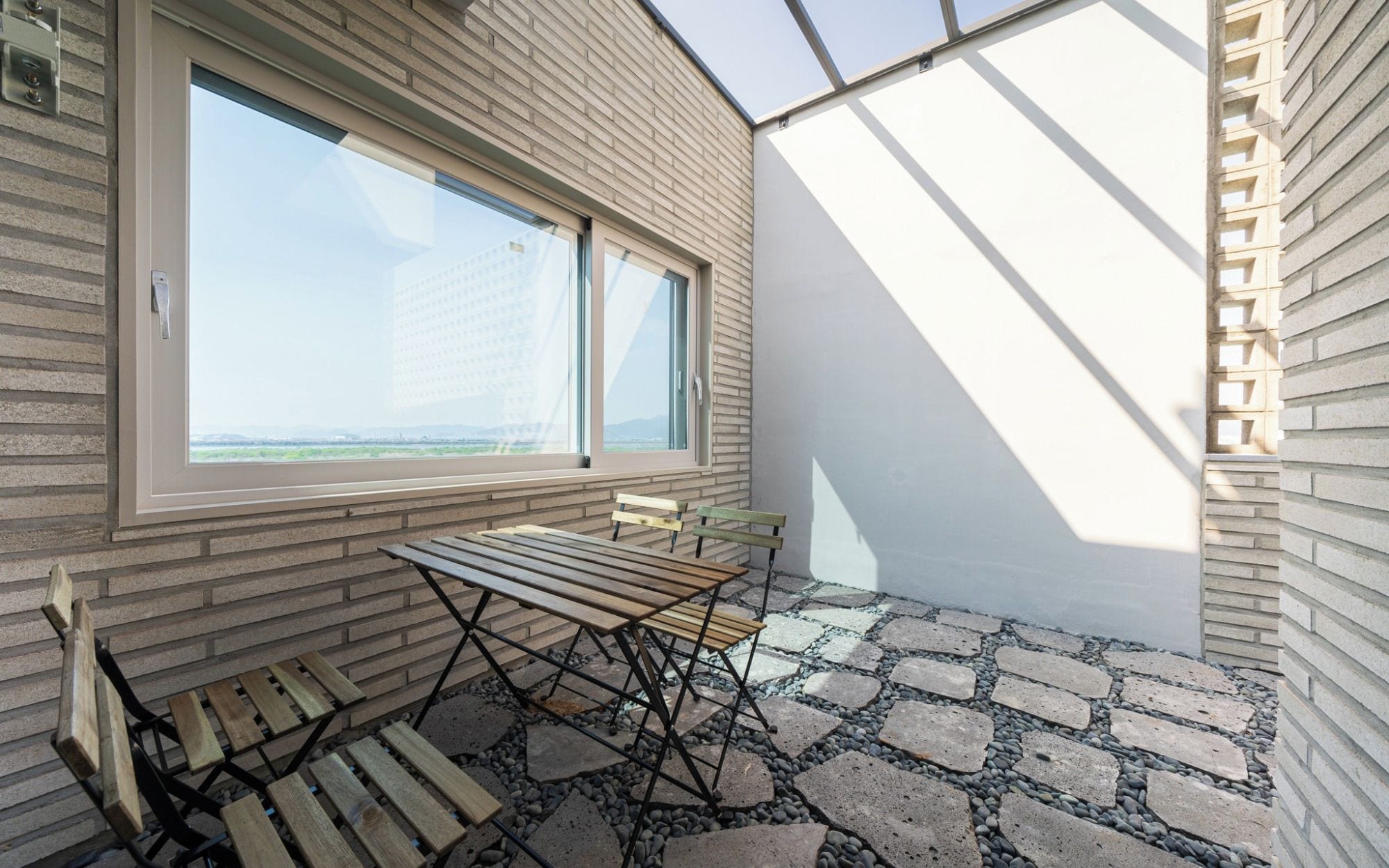 A balcony with a table and benches, a large window with a view, and a patterned stone floor.