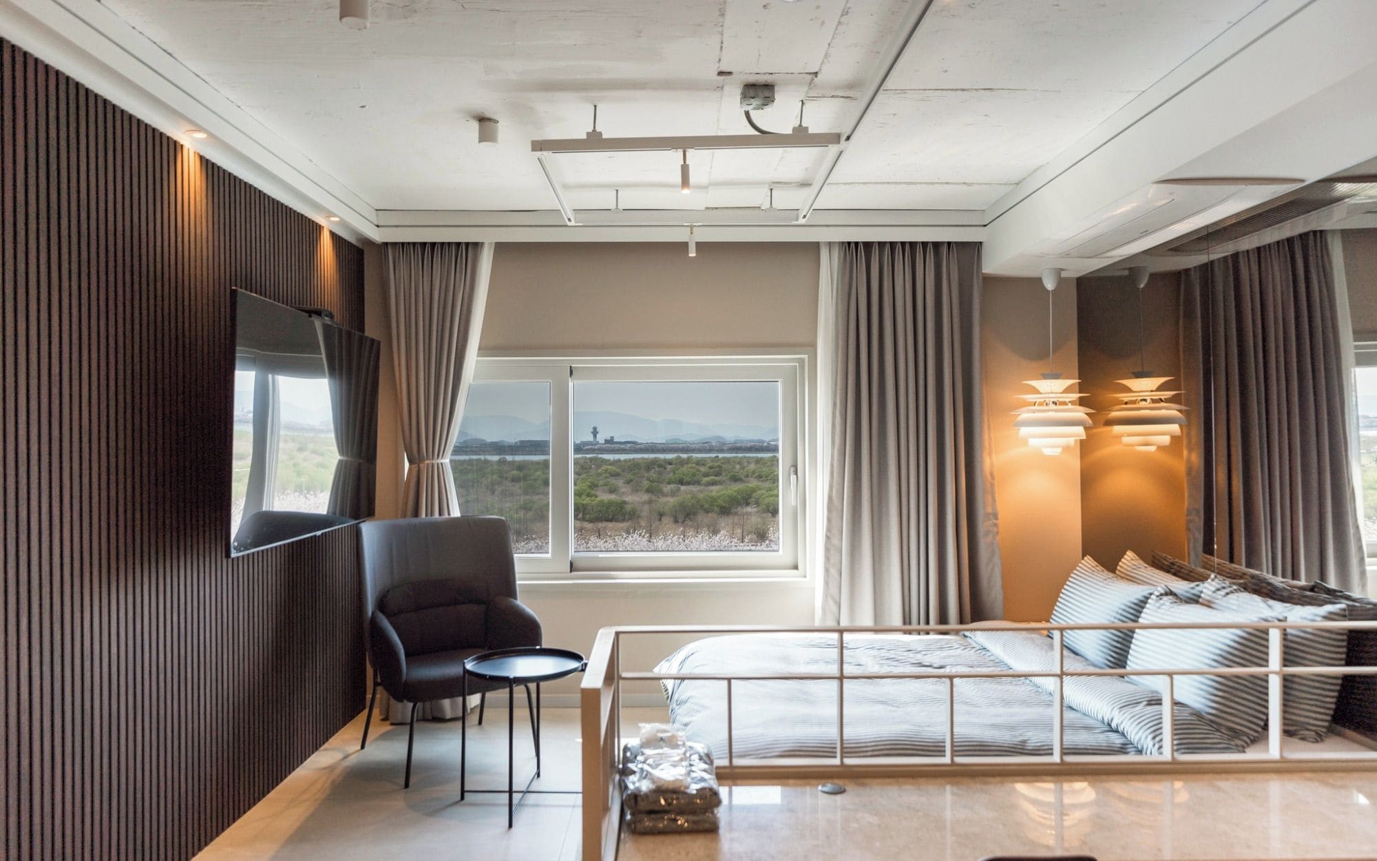 A modern hotel room with a bed, a chair, a small table, a TV mounted on the wall, and a large window offering a view of the landscape.