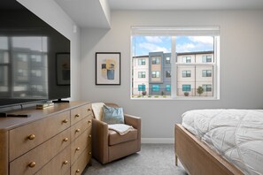 A bedroom with a bed, a dresser, a chair, a television, and a window with a view of buildings. - Splash Zone | A Family Favorite near Zion (Hurricane)