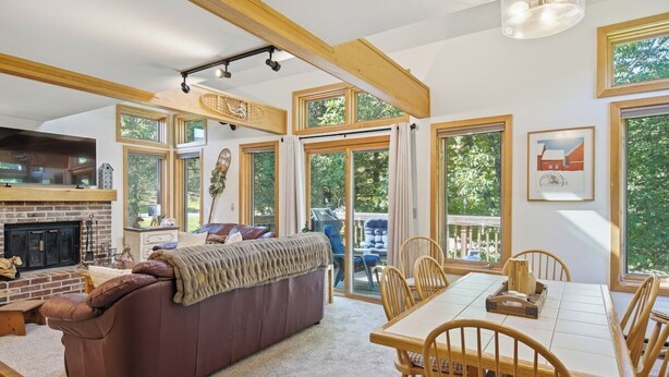 A spacious living area with a fireplace, a large sofa, a dining table with chairs, and large windows offering a view of the outdoors. - The Back Nine | Golf, Mount Snow, & Vermont Escape (Wilmington)