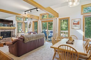 A spacious living area with a fireplace, a large sofa, a dining table with chairs, and large windows offering a view of the outdoors. - The Back Nine | Golf, Mount Snow, & Vermont Escape (Wilmington)