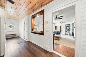 En gang med træloft, en bænk, et spejl og udsigt til en opholdsstue med en blå sofa og en ventilator.
