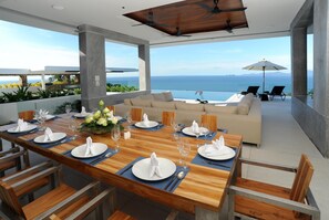 Un espace repas moderne avec une table en bois dressée pour le repas, offrant une vue panoramique sur l’océan.