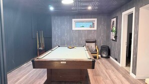 A pool table with a billiard cue rack, a single chair, and a speaker in a room with a wooden floor and a window with a view. - Zen on the Lake Spa | Mission, BC (Mission)