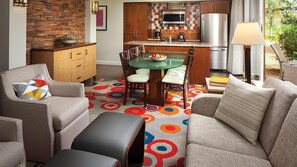 A modern kitchen with a stone backsplash, wooden cabinets, and stainless steel appliances. There is a dining table with chairs, a sofa, and a colorful circular rug. - Prime location for festivals, golf, and restaurants w/2 full bedrooms & baths (Palm Desert)