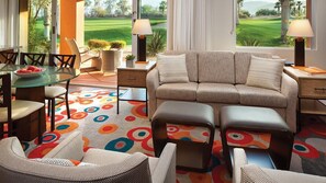 A living room with a sofa, ottoman, and chairs, a glass table with a bowl of fruit, and a view of a golf course through large windows. - Prime location for festivals, golf, and restaurants w/2 full bedrooms & baths (Palm Desert)