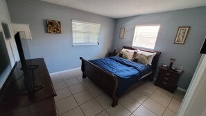 A bedroom with a bed, a dresser, two bedside tables, a window with blinds, and a wall-mounted TV. - You'll love it here (Palm Beach Gardens)