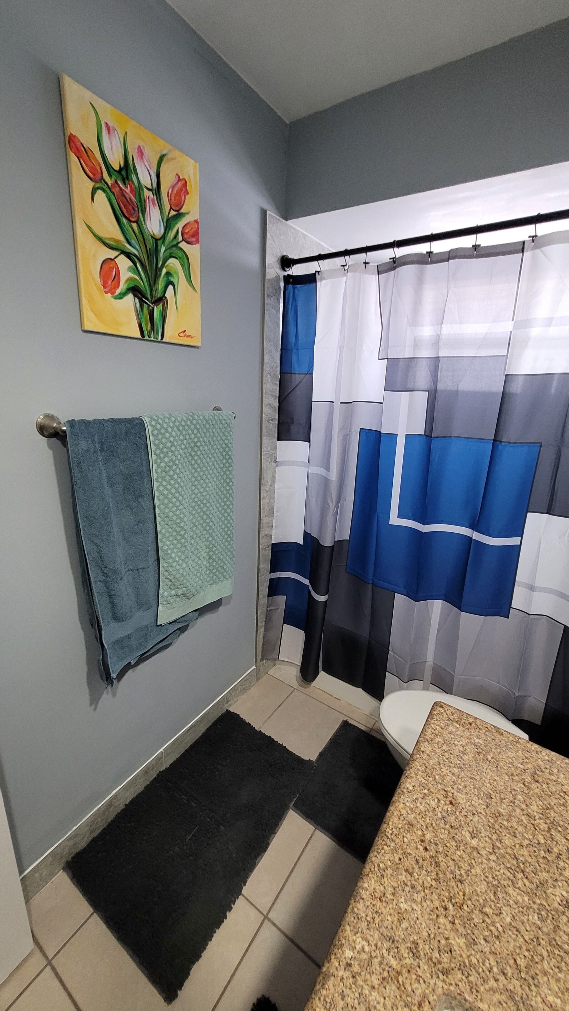 A bathroom with a shower curtain, a towel rack with a towel, a painting on the wall, and a tiled floor.