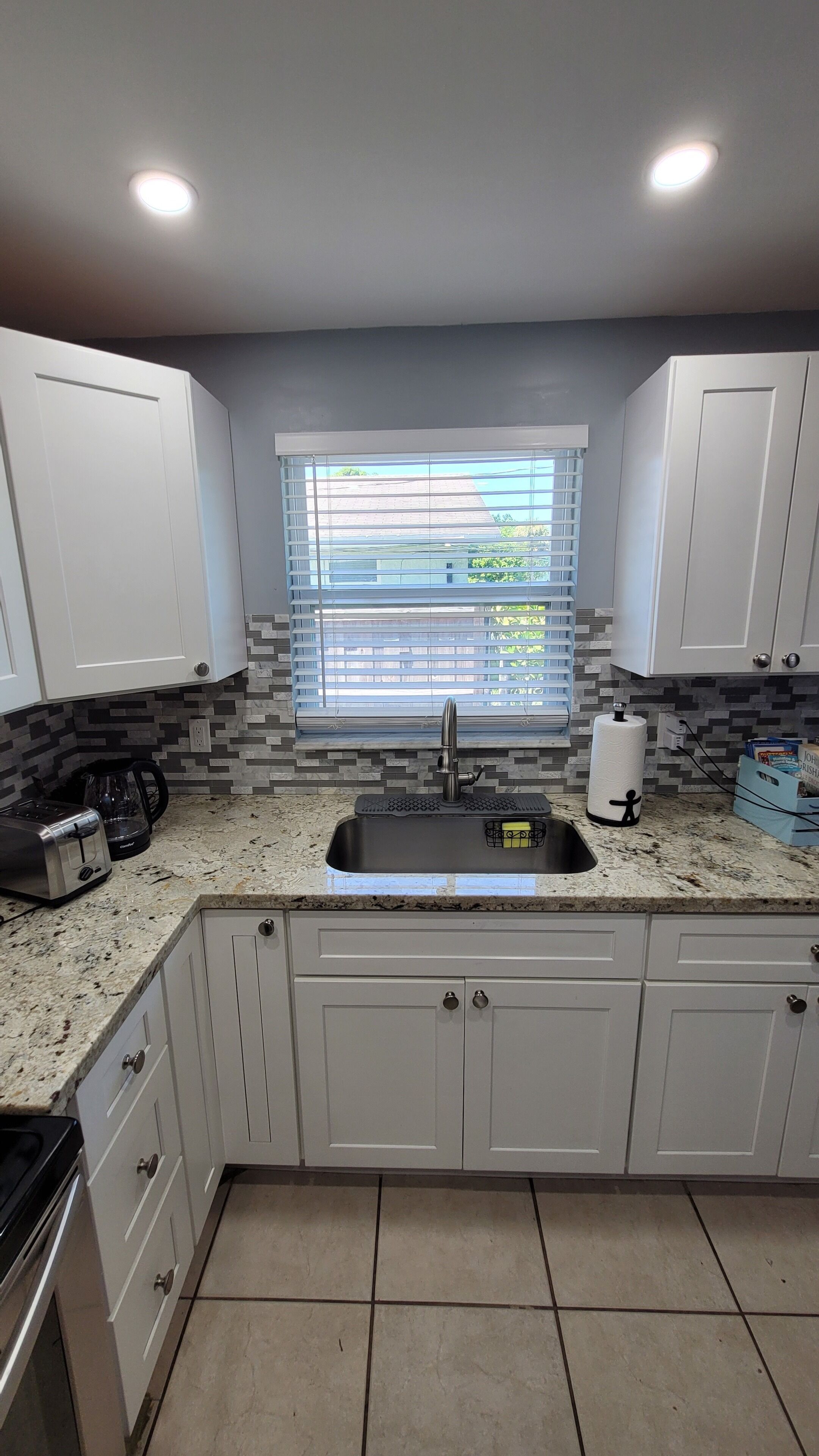 A kitchen with white cabinets, a granite countertop, a window with blinds, and a stainless steel sink.