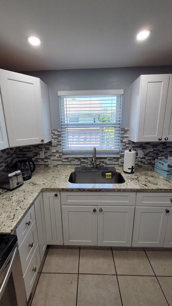 A kitchen with white cabinets, a granite countertop, a window with blinds, and a stainless steel sink.