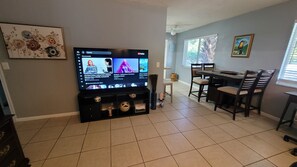 A living room with a flat-screen TV displaying a music video, a dining area with a table and chairs, and a painting on the wall.