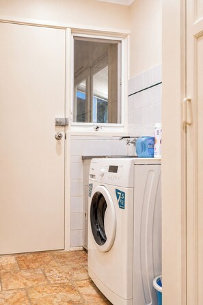 A compact laundry room with a front-loading washing machine, a small window, and a tiled backsplash. - Relaxing 4BR Wantirna Home | Everything You Need (Wantirna South)