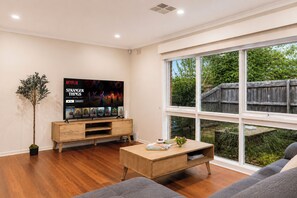 A modern living room with a large TV, wooden furniture, and a view of the outdoors. - Relaxing 4BR Wantirna Home | Everything You Need (Wantirna South)