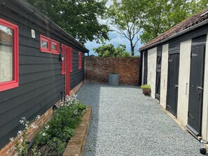Ein Innenhof mit Kiesweg, einer Backsteinmauer und zwei Gebäuden mit roten Fensterrahmen und schwarzen Türen.