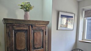 Ett träskåp med en vas med blommor ovanpå, en inramad tavla på väggen och ett fönster med utsikt.