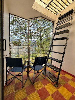 Une pièce d’angle donnant sur une cour grâce à une grille en métal, avec deux chaises, une échelle et un sol à carreaux.