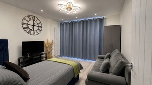 A modern bedroom with a bed, a sofa, a TV, a decorative clock, and a ceiling light fixture. - House Sleeps 9 with Wifi (Slough)