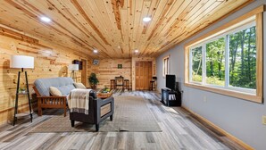 Ett mysigt vardagsrum med en soffa, ett soffbord och en tv, träpanel på väggarna och ett stort fönster med utsikt över träd.