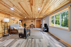 Ein gemütliches Wohnzimmer mit einer Couch, einem Couchtisch und einem Fernseher, Holzwänden und einem großen Fenster mit Blick auf Bäume.
