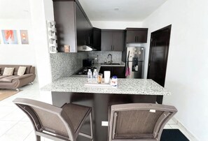 A compact kitchen with dark cabinets, a granite countertop, a refrigerator, and a dining area with two chairs.