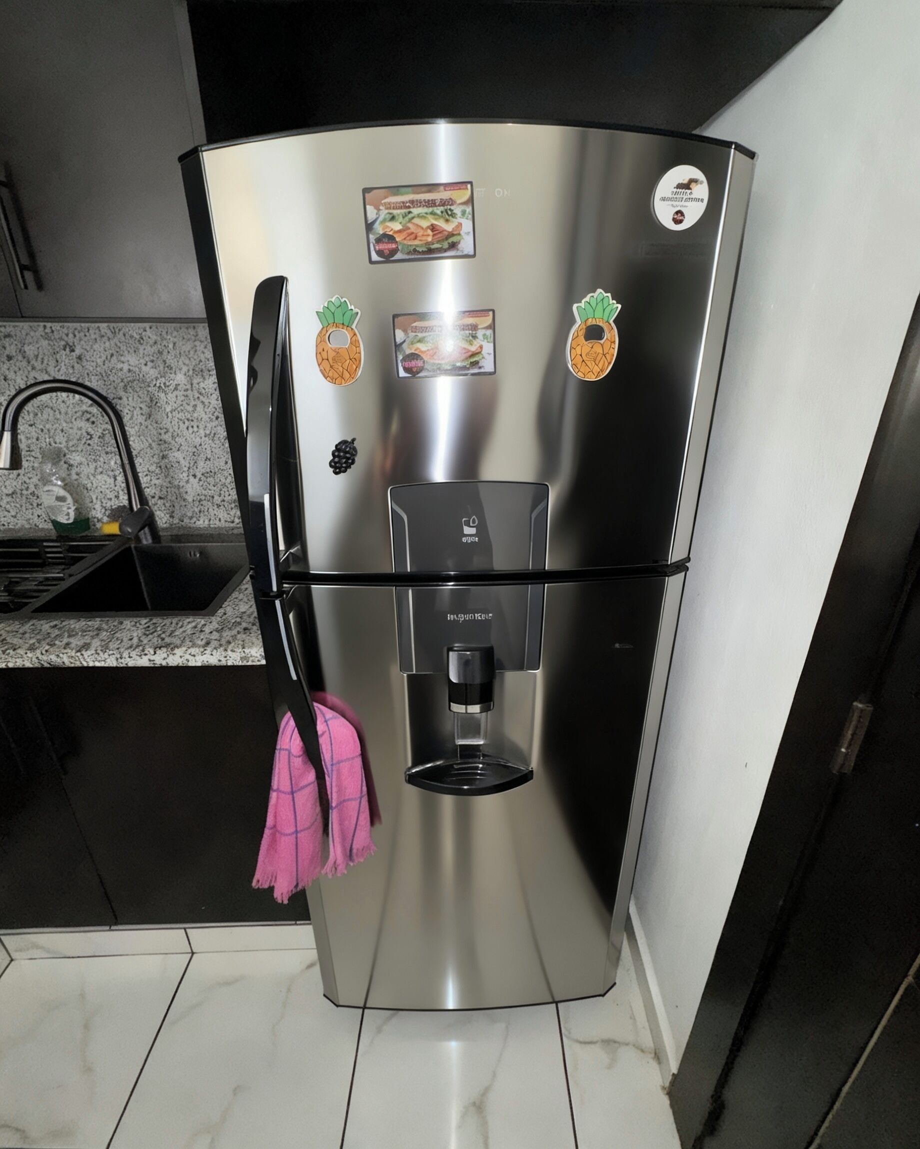 Frigorifero in acciaio inox con erogatore d'acqua, due calamite e un asciugamano rosa appeso alla maniglia.