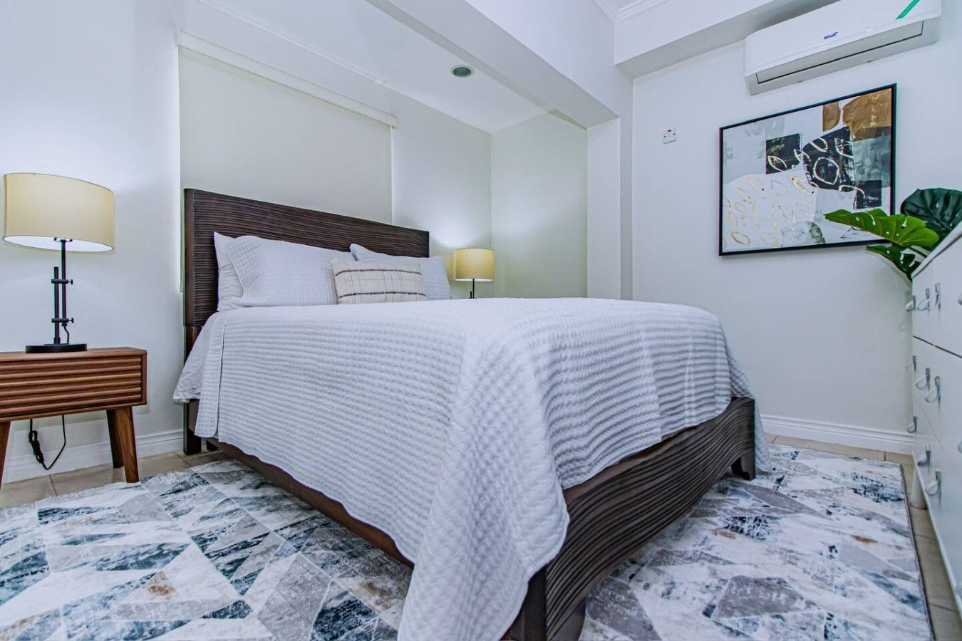 Een slaapkamer met een bed, nachtlampjes, een houten nachtkastje, een vloer met een marmerlook en een wandairconditioning.