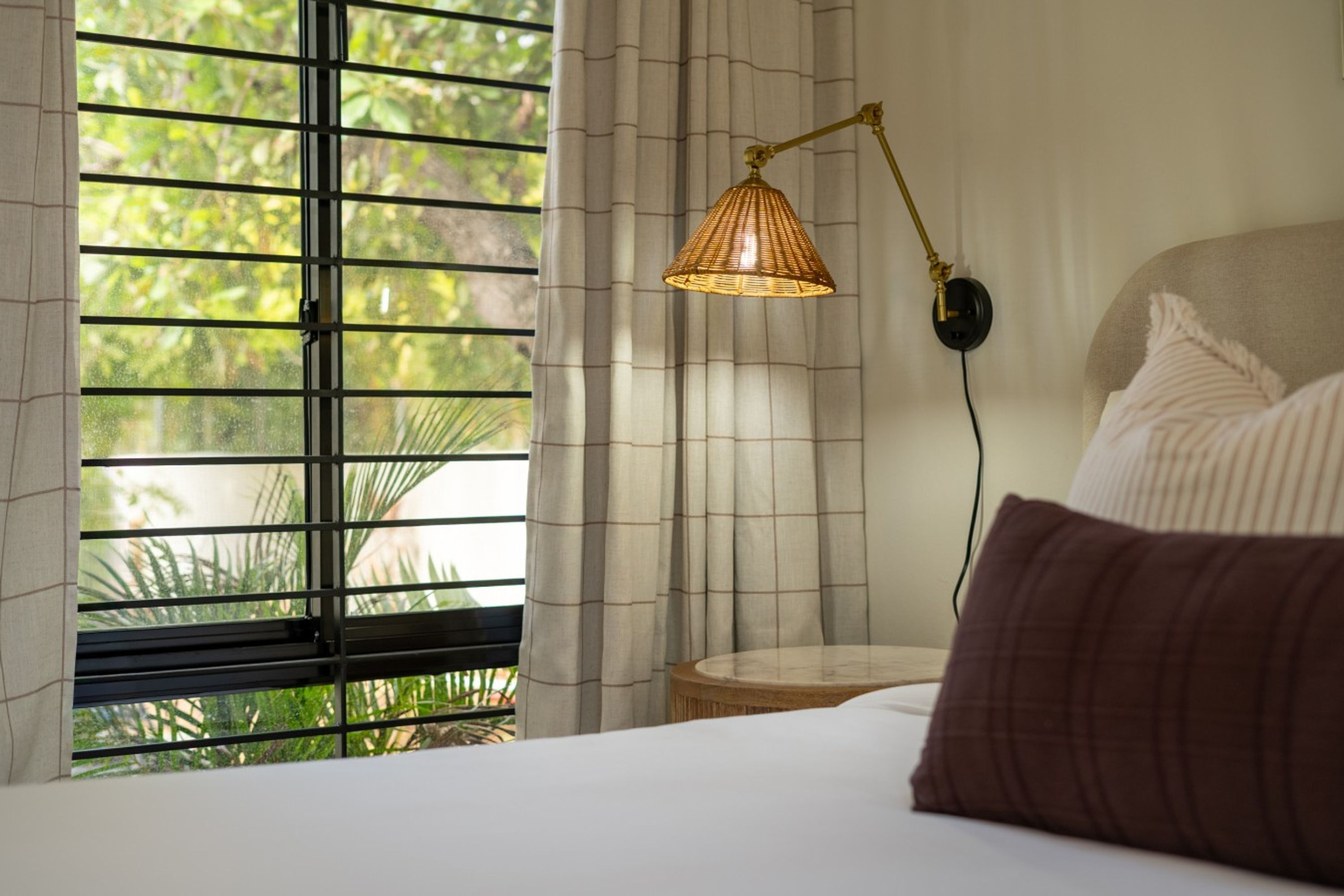 Quarto com cama, luminária de cabeceira, janela com persianas e vista para o verde lá fora.