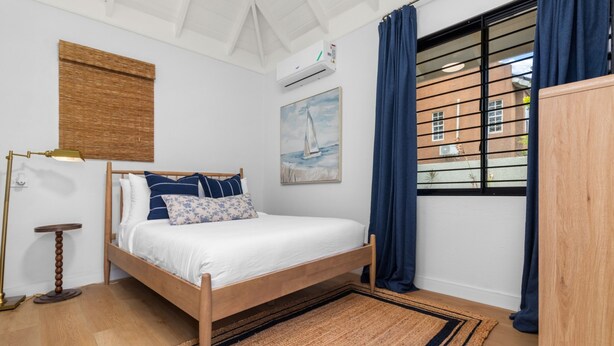 A bedroom with a wooden bed, a bedside lamp, a rug, a painting, and a window with curtains. - 5BR Luxe Retreat in Runaway Bay Private Pool Beach Access (Runaway Bay)
