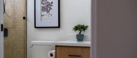 A bathroom with a toilet, a framed botanical illustration, and a small potted plant on the counter.