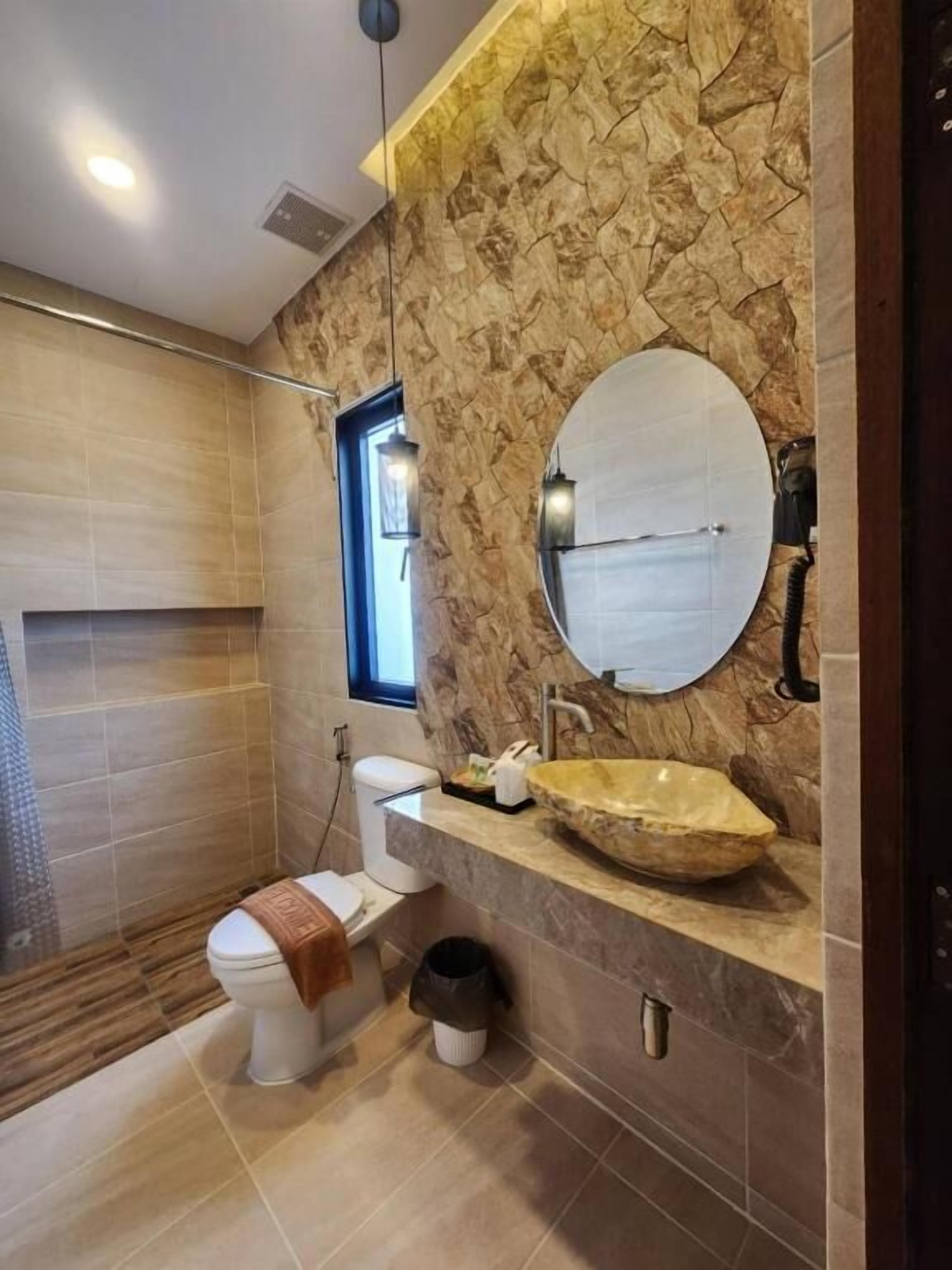 A modern bathroom with a stone wall, a round mirror, a vessel sink, and a toilet.