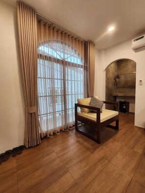 A room with a wooden floor, a bed with a pillow, a window with curtains, and a wall-mounted air conditioner. - Baankorrakang (Bangkok)