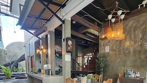 A restaurant interior with a central dining table, wooden chairs, hanging pendant lights, and a staircase leading to an upper level.