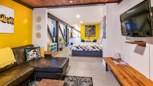 A modern living space with a sofa, a wooden table, a TV mounted on the wall, and a bed with a patterned bedspread.