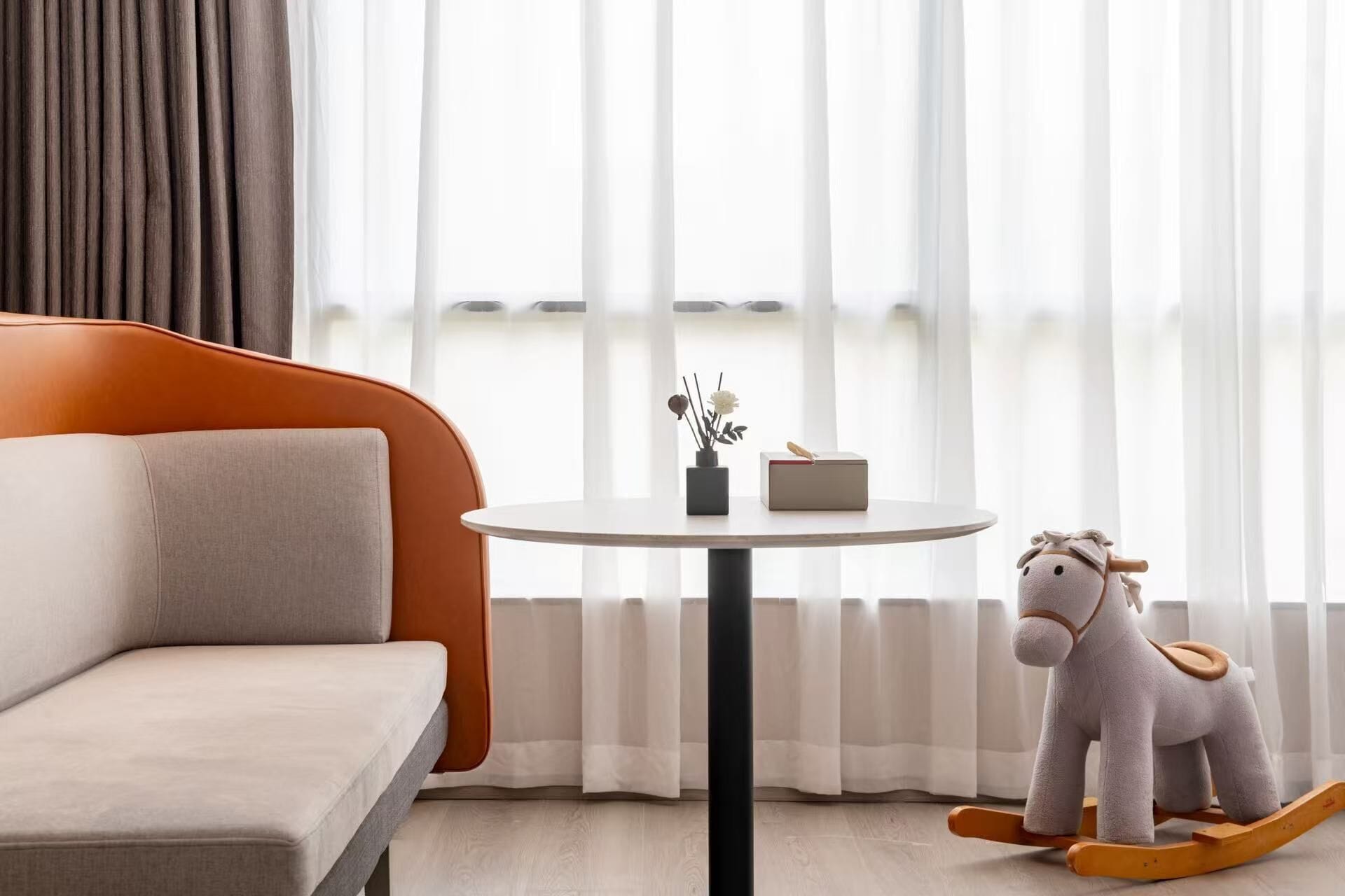 Una sala de estar moderna con un sofá, una mesa redonda, un caballito de madera y una ventana con cortinas translúcidas.