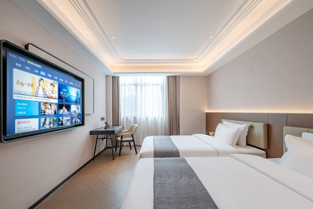 A hotel room with a large flat-screen TV mounted on the wall, a desk with a chair, two beds, and a window with curtains.