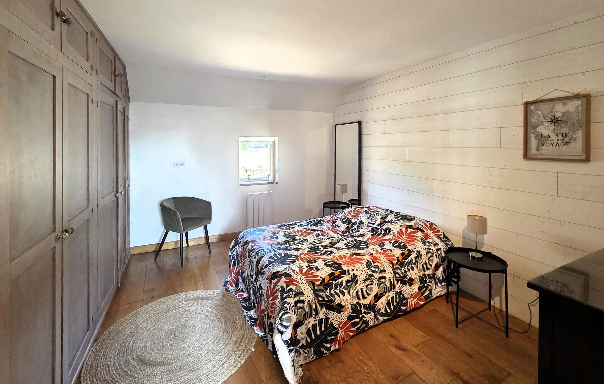 A bedroom with a bed, a chair, a small table, a mirror, and a framed picture on the wall.
