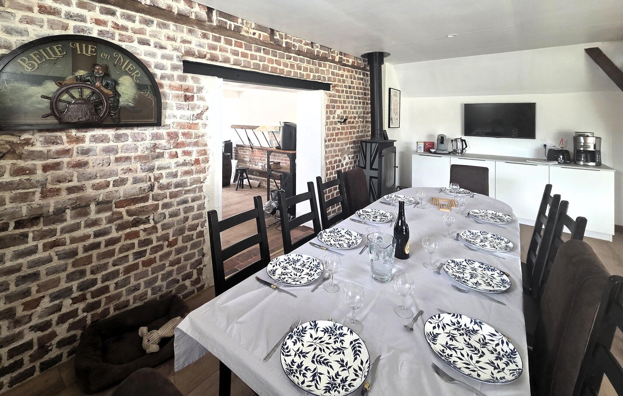 A dining area with a brick wall, a table set for six with plates, glasses, and cutlery, and a television mounted on the wall.