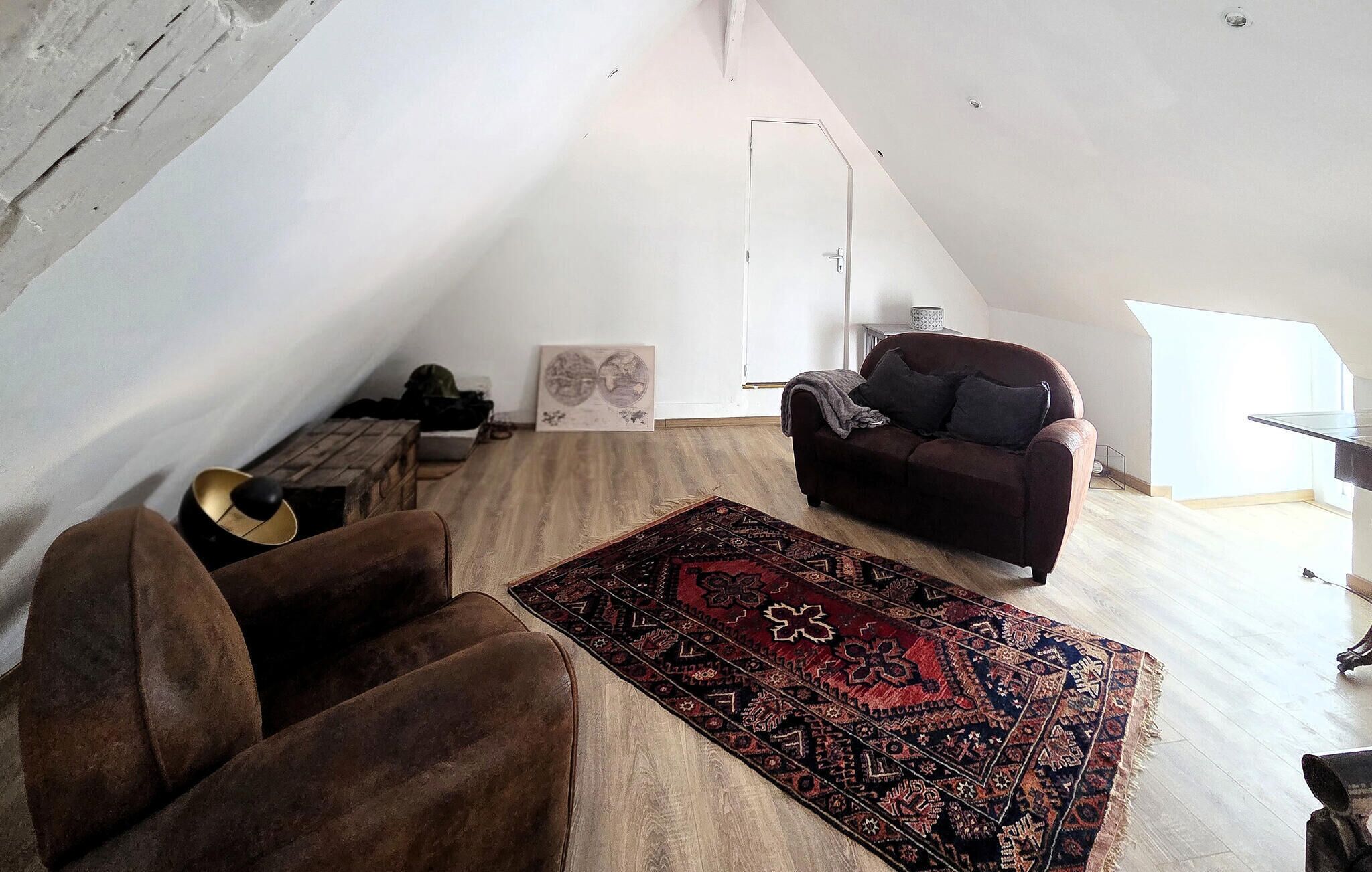 A loft-style room with a sloped ceiling, a brown leather sofa, a patterned rug, and a dark brown armchair.