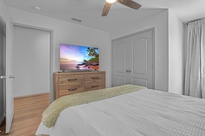 A bedroom with a bed, a wooden dresser, a television on the wall, and a ceiling fan.
