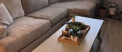 A living room with a beige sectional sofa, a wooden coffee table with plants, a ceiling fan, and a window with blinds.