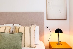 A neatly made bed with striped pillows, a plain wall with a framed artwork, and a bedside table with a lamp. - Pink Calas Blancas Apartment (Torrevieja)